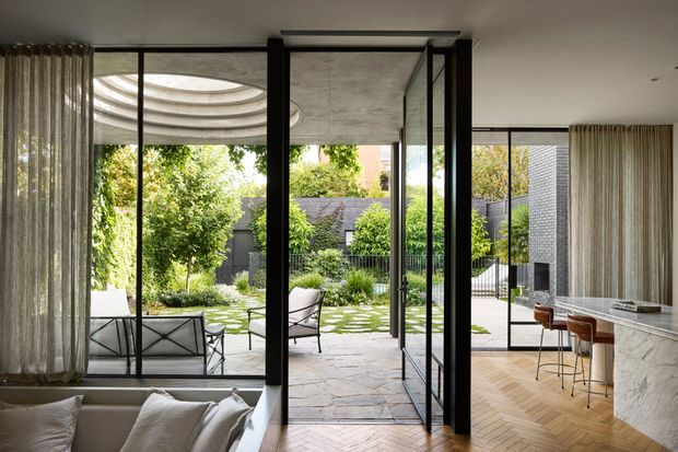 A refined contemporary open-plan living and kitchen space opening through large black-framed glazed doors onto a landscaped courtyard. The room features a pale marble kitchen island, timber herringbone flooring, neutral upholstered seating, sheer curtains, and a partially visible sculptural ceiling detail. The courtyard includes paved stone, greenery, outdoor seating, and a dark masonry boundary wall.