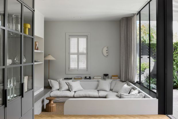 Refined contemporary living room with a large light-grey sectional sofa integrated into a raised, white-edged seating platform. The room features full-height black-framed glazing opening toward a planted outdoor area, a central shuttered window, full-length taupe curtains, built-in grey cabinetry with glass doors, a sculptural wall light, and pale herringbone timber flooring. The palette is soft and neutral with strong black-framed glazing accents.