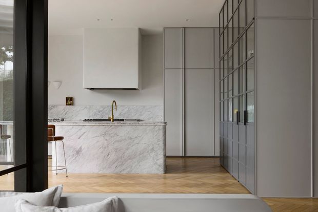 A refined open-plan kitchen viewed from an adjacent living area. The space features a large white marble island with waterfall sides, a brass tap, pale integrated cabinetry, herringbone timber flooring, and a full-height black-framed glazed partition enclosing the kitchen or adjoining room. Neutral finishes and concealed appliances create a clean, sophisticated interior.