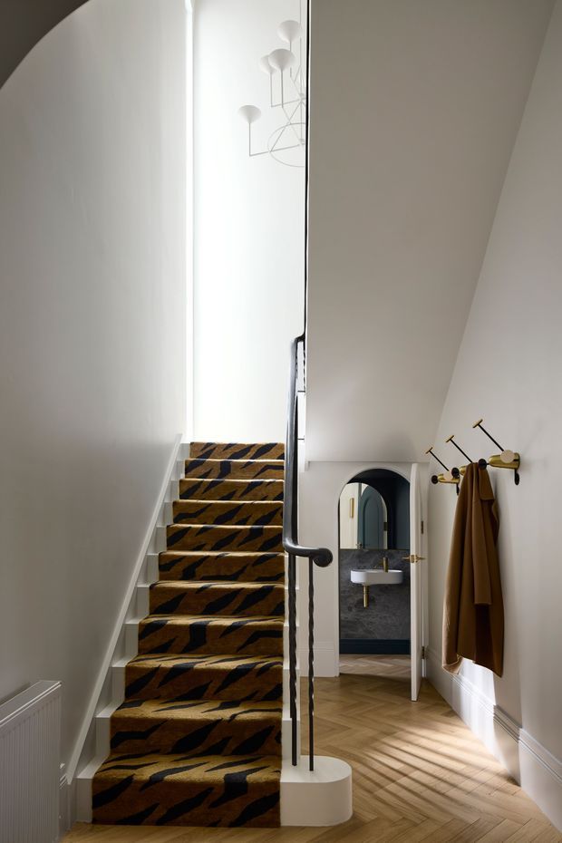 A refined residential stairway and hallway with a sculptural white staircase, dark handrail, and bold patterned stair runner. White walls and decorative moulding contrast with warm herringbone timber flooring. An arched doorway leads to a compact bathroom with a wall-mounted basin, while a minimalist brass-and-black coat rack and contemporary pendant lights add character.