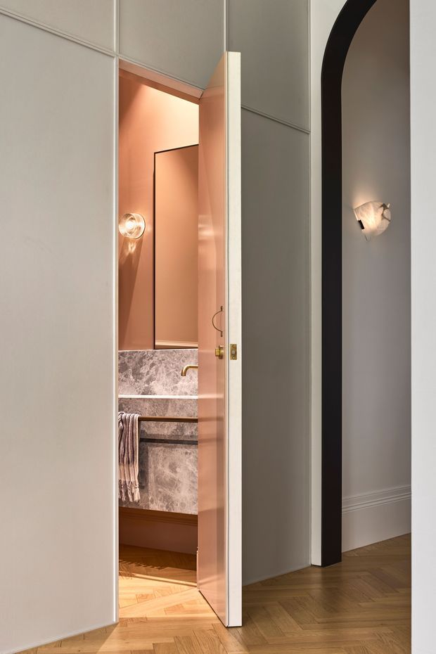 A refined interior view looking toward a partially open bathroom door. The bathroom has warm peach-toned walls, a marble-look vanity or wall surface, a brass tap, a round wall light, and a striped hand towel. The surrounding hallway features pale grey wall panelling with moulded trim, a dramatic black arched opening, warm herringbone timber flooring, and a sculptural wall sconce. The atmosphere is warm, elegant, and quietly luxurious.