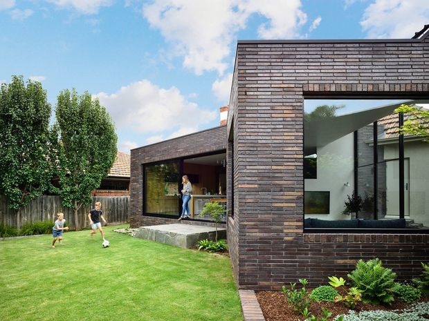 Exterior view of a contemporary single-storey brick-clad home with strong rectilinear volumes and large black-framed glazed openings. A covered outdoor living area connects to the lawn and garden, while the interior is partially visible through the glazing. The landscaped suburban setting includes a family lawn and planted garden beds under a bright sky.