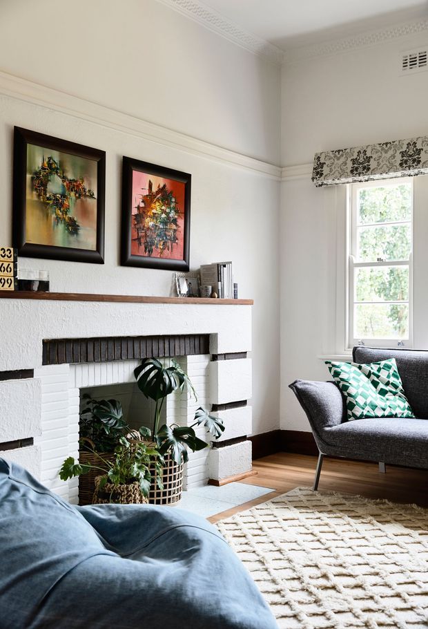 Bright, refined living room with a white painted brick fireplace, timber mantel shelf, dark timber skirting, hardwood flooring, framed artwork, a grey upholstered armchair, blue sofa, patterned cushions, indoor plants, and a textured cream rug. The room features decorative ceiling cornices and a patterned Roman blind, with daylight entering through a tall sash-style window.