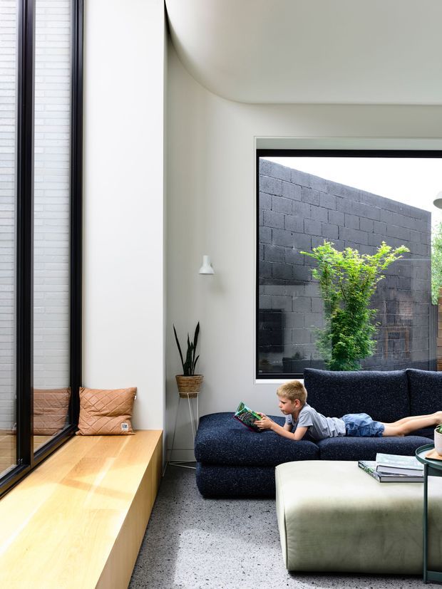 A bright, contemporary living room with a large dark upholstered sectional sofa and ottoman, a child reading, and a built-in light timber window seat beside tall black-framed glazing. The room features a softly curved white ceiling transition, pale walls, a speckled grey floor, and a large window looking onto a planted courtyard enclosed by a dark concrete-block wall.