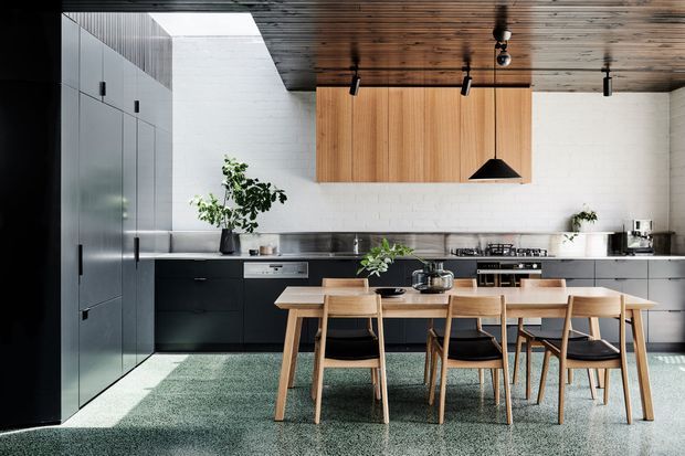 A spacious contemporary kitchen and dining area with full-height matte dark cabinetry, a long dark benchtop, white brick-look wall finish, warm timber overhead cabinetry, and a timber slatted ceiling. A large rectangular dining table with timber chairs occupies the foreground, while black track lighting and a sculptural black pendant provide contrast. The space has a refined industrial-modern character with abundant natural light.