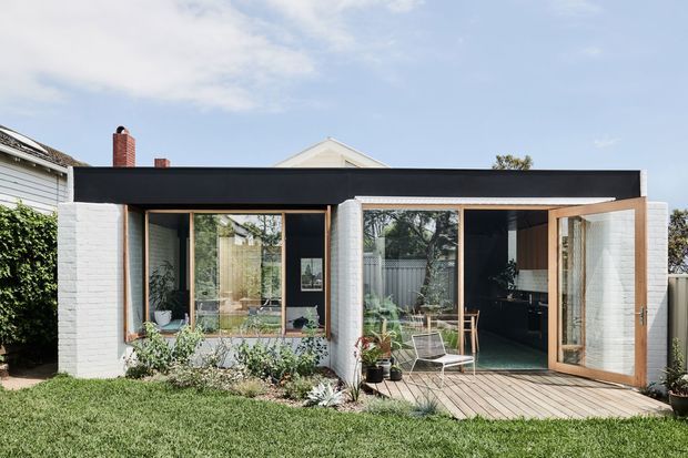 A small modern single-storey extension or pavilion with a bold flat black roof, white painted brick walls, warm timber-framed glazing, and large sliding or folding glass doors opening onto a timber deck. The structure sits in a landscaped suburban garden and includes a dark built-in interior area visible through the glazing.
