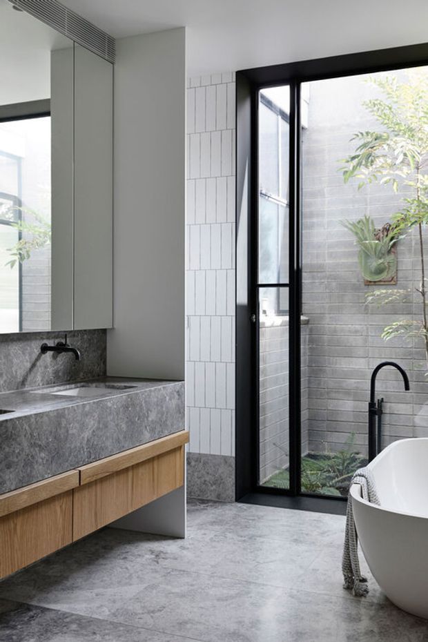 Bright contemporary bathroom featuring a floating timber vanity with a grey stone benchtop and integrated basin, black tapware, a freestanding white bathtub, large-format grey stone-look flooring, and a black-framed glazed door opening to a planted courtyard. A vertical white tile feature wall and grey masonry courtyard wall add texture.