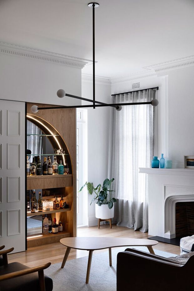 A refined living room in a heritage-style residence, featuring ornate white ceiling cornices and wall detailing, tall sheer-curtained windows, and a white traditional fireplace. A built-in timber drinks cabinet with an illuminated arched display and glass shelving occupies the left wall. Mid-century-inspired dark leather seating and a rounded timber coffee table sit on a pale textured rug, beneath a striking black linear multi-arm pendant light. The palette is predominantly white, warm timber, black, and muted blue accents.