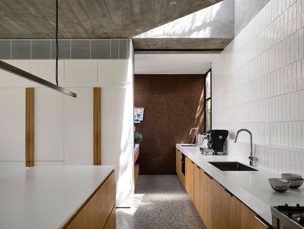 Contemporary galley kitchen with a long white benchtop, timber cabinetry, integrated sink and cooktop, and a tiled white wall. The space features exposed concrete ceiling planes, a skylight opening, terrazzo-style flooring, and a dark textured feature wall at the rear. A pendant light and partial island cabinetry are visible.