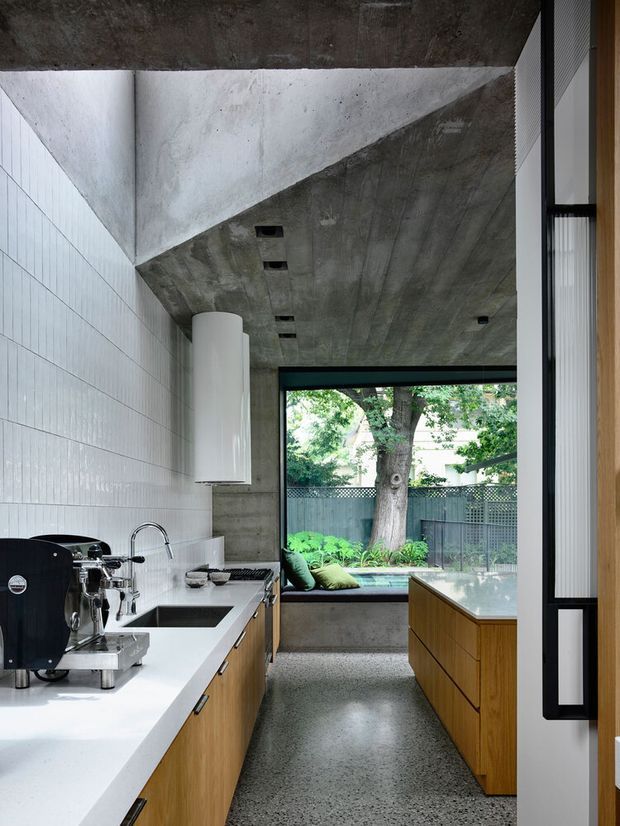 A narrow contemporary kitchen with a concrete ceiling and structural elements, white vertically tiled walls, pale stone benchtops, and warm timber cabinetry. A large black-framed window looks onto a leafy courtyard, while a built-in window seat with cushions sits at the rear. The kitchen includes a coffee machine, sink, concealed appliances, and a central island with a pale benchtop.