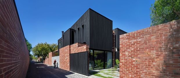 Exterior view of a contemporary multi-level residential building in a narrow urban lane. The design combines dark vertical cladding, exposed red brick walls, large glazed openings, and strong angular roof volumes, with a small landscaped courtyard visible at the entrance.