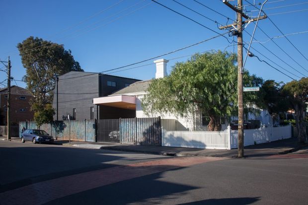 Street-level exterior view of a suburban residential property at a street corner. The foreground shows asphalt roads, a red-paved footpath, utility poles and overhead wires. The property includes a contemporary dark-clad addition with a projecting flat canopy, alongside a lighter older dwelling with a pitched roof, chimney, front fence and mature trees.