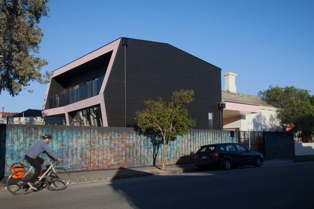 Street view of a contemporary two-storey building with a sharply folded geometric facade, dark horizontal cladding, and contrasting pale pink rendered structural surfaces around recessed glazing and balconies. A weathered painted fence, mature trees, neighbouring homes, parked car, and cyclist occupy the foreground and surroundings.