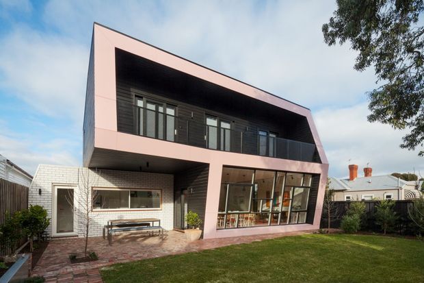 Exterior view of a contemporary two-storey residence with a dramatic cantilevered upper level and large angular pink-painted frame. The lower level combines white brickwork, extensive glazing, and a sheltered patio, while the upper level has dark horizontal cladding and a recessed balcony or terrace. A lawn and paved path occupy the foreground, with neighbouring homes visible beyond.