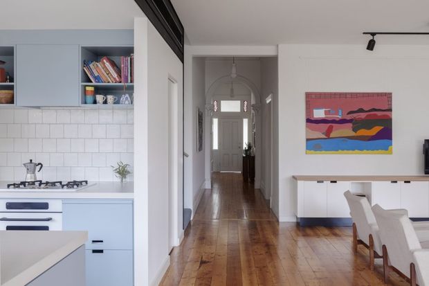 Bright open-plan residential interior showing a light blue and white kitchen on the left, a timber-floored hallway leading to the front door in the centre, and a dining/living area with white built-in cabinetry and dining chairs on the right. A colourful abstract artwork and black track lighting add visual accents. The space has a clean, contemporary feel with timber flooring and restrained built-in joinery.