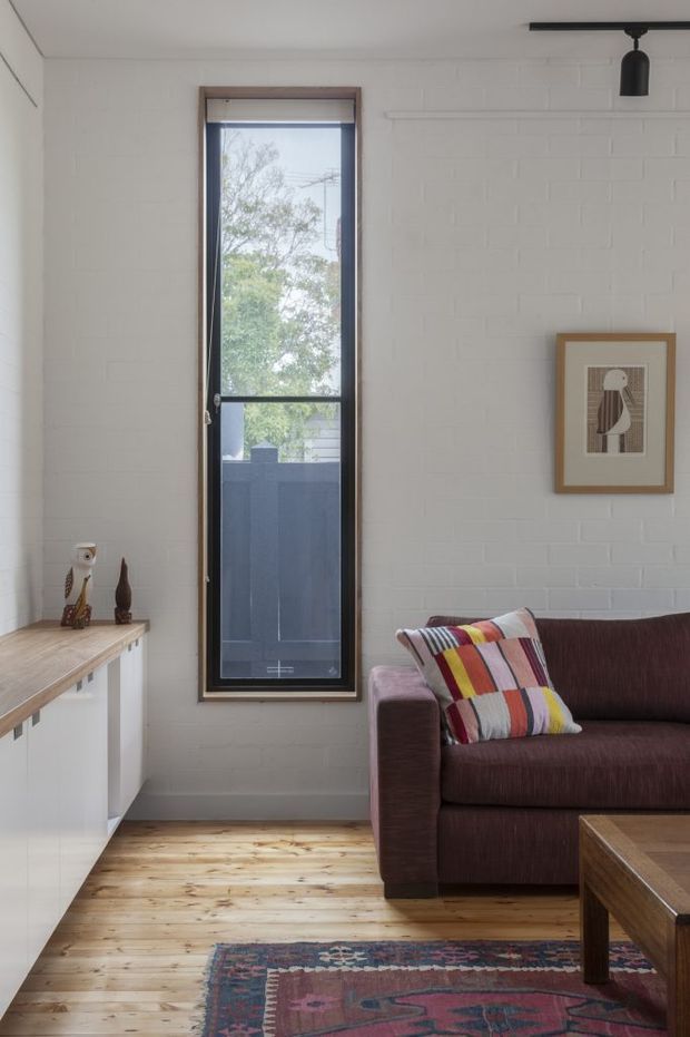 A bright, compact living room with white-painted brick walls, warm timber floorboards, and a tall narrow window with dark framing. A deep burgundy sofa with a colourful striped cushion sits beside a timber coffee table, while a low built-in cabinet and framed artwork add simple character. A black ceiling-mounted spotlight provides focused lighting.