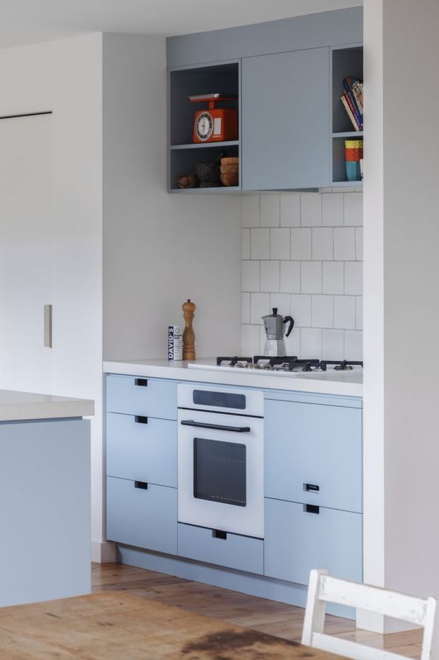A compact, softly lit kitchen alcove with pale blue flat-panel cabinetry, black pulls, a white tiled subway backsplash, white benchtop, and a built-in oven with gas cooktop. Open upper shelving displays books and pottery, while a timber dining table and white chair are visible in the foreground.
