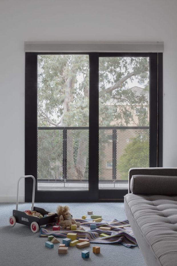A bright, minimalist living room viewed toward full-height black-framed glazed doors opening onto a balcony with perforated metal screening. A large grey upholstered sofa occupies the foreground, while a children's play wagon, soft toys, colourful wooden blocks and a knitted play blanket sit on the pale carpet. White walls and simple roller blinds create a clean, contemporary interior.