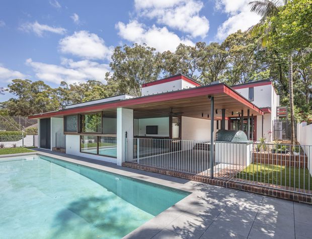 Exterior view of a low-slung modernist house with strong mid-century influences, featuring broad roof overhangs, a red-edged flat roof, extensive glazing, white rendered walls, and a covered outdoor entertaining area. A swimming pool and paved terrace occupy the foreground, with landscaped planting and mature trees surrounding the property.
