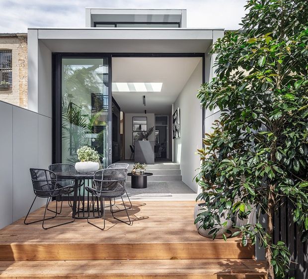 A contemporary urban townhouse extension viewed from a timber deck, with a compact outdoor dining setting in the foreground and large dark-framed glazing opening into a bright, minimalist interior. The flat-roofed addition has a roof-level clerestory or raised glazed element, a skylight over the entry, crisp white walls, and a refined indoor-outdoor connection. Mature leafy planting screens the right side of the deck.