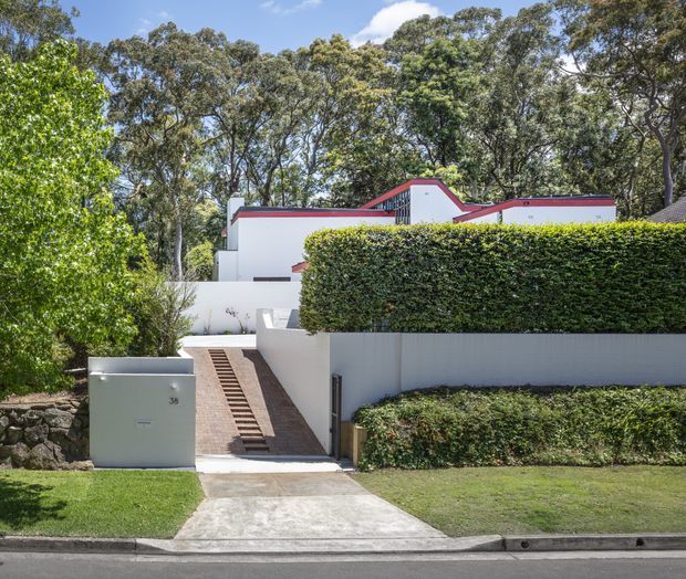 Street-facing view of a modern white rendered residence set within mature leafy landscaping. The property has a low, flat roof profile with distinctive red metal roof-edge trims, tall manicured hedges, rendered boundary walls, a sloping paved pedestrian entry with steps, and a concrete driveway or path leading from the footpath.