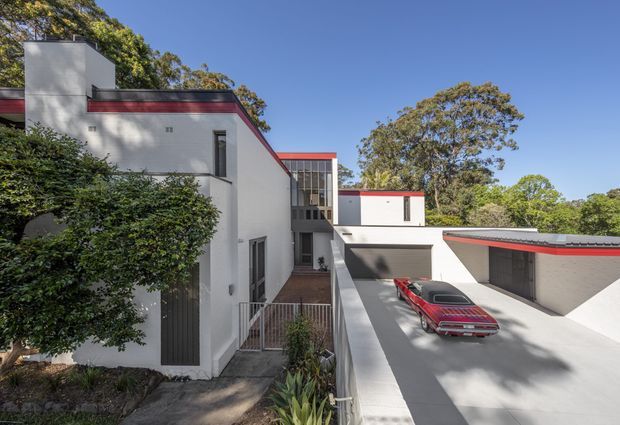 Exterior view of a contemporary multi-level residence set among mature trees. The building has crisp white rendered walls, strong horizontal roof and parapet lines with dark grey roofing and red accent trim, tall glazed stairwell or circulation windows, and an integrated covered garage or carport area. A red vintage car is parked on the concrete driveway, while landscaping frames the left side of the property.