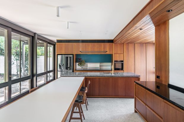 A warm contemporary kitchen with extensive walnut-toned timber cabinetry and a matching timber-lined ceiling. The room features a long white island with bar seating, a dark stone kitchen benchtop, integrated appliances, black-framed floor-to-ceiling windows, and a terrazzo-like light floor. Abundant glazing brings in greenery and daylight, creating a refined open-plan atmosphere.
