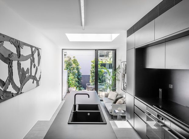 A bright, narrow contemporary kitchen opening toward a glazed outdoor courtyard. The room features a long dark island with an undermount double-bowl sink and black tap, extensive full-height dark cabinetry with integrated appliances, pale tiled flooring, white walls, a linear ceiling vent, and a roof skylight. A compact living area with seating is visible beyond the kitchen, along with greenery through the glazing. A large monochrome abstract artwork occupies the left wall.