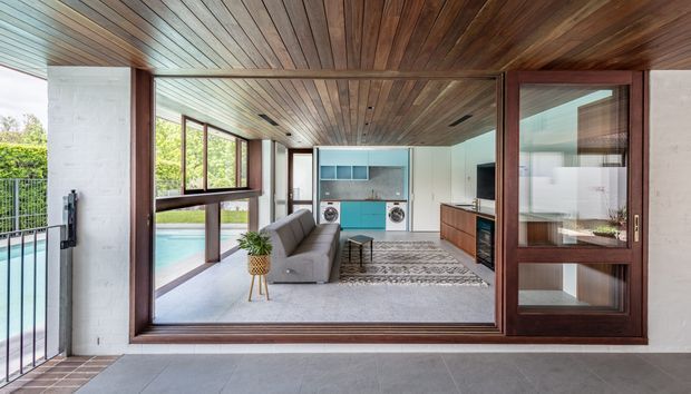 A bright contemporary open-plan living area viewed through large timber-framed glazed doors. The room features a grey sofa, patterned rug, dark timber built-in cabinetry, and a compact turquoise-accented kitchen at the rear. Warm timber lining covers the ceiling, while expansive glazing opens toward a swimming pool and leafy outdoor setting.
