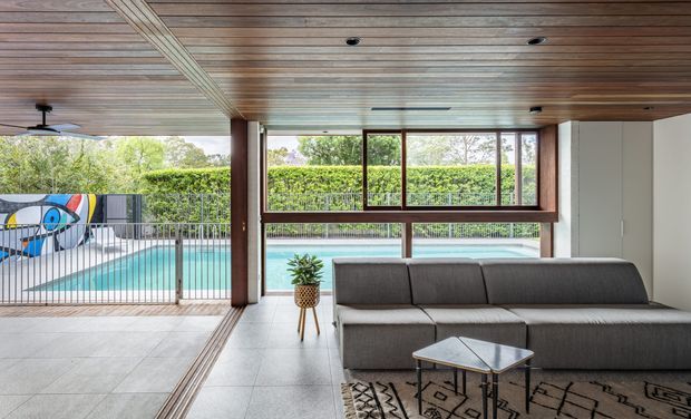 A covered indoor-outdoor living area overlooking a swimming pool. The space features a large dark sectional sofa, patterned rug, small coffee table, tiled flooring, exposed timber ceiling lining, timber-framed glazing, and wide openings to the pool terrace. A colourful mural is visible beyond the metal balustrade.