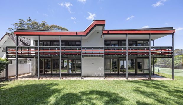 Front exterior view of a two-storey contemporary building with a strong horizontal composition, dark framed glazing, extensive upper-level balconies, and a distinctive red-trimmed stepped roofline. The facade combines light rendered walls, dark metal framing, and transparent ground-floor glazing, set beside a neatly maintained lawn and outdoor sports court.