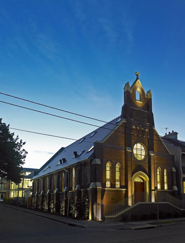 Exterior architectural photograph of a restored or repurposed Gothic Revival brick church in an urban streetscape at dusk. The building has a steeply pitched metal roof, tall central bell tower, pointed-arch openings, round traceried window, decorative brickwork, illuminated arched windows, and a stepped accessible entrance. Overhead utility wires and neighbouring buildings are visible.