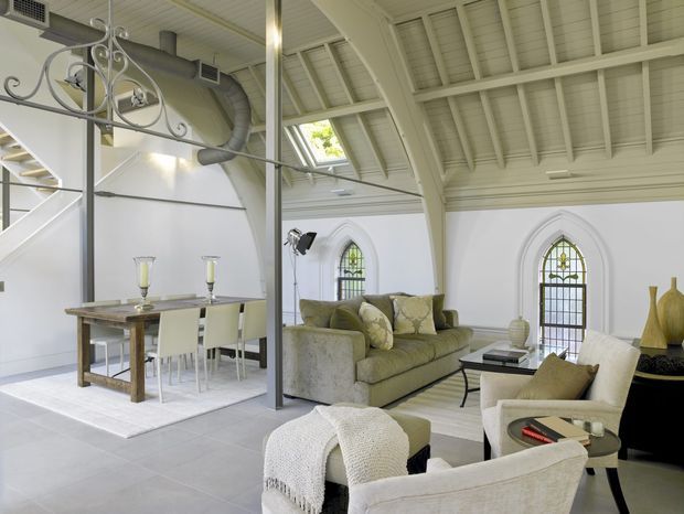 Bright open-plan living and dining space in a converted chapel-like interior, featuring a high exposed pitched timber roof with painted rafters, tall pointed-arch stained-glass windows, white walls, pale tiled flooring, and a decorative metal room divider. A neutral upholstered sofa and armchairs form the living area beside a timber dining table and white dining chairs.