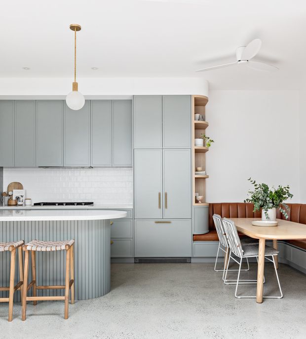 Bright open-plan kitchen and dining nook with pale blue-grey panelled cabinetry, a curved island with vertical fluted detailing, white subway-tile splashback, integrated appliances, and warm timber open shelving. A built-in cognac leather banquette wraps around a light timber dining table with white metal chairs. The room has a smooth white ceiling, contemporary pendant lighting, ceiling fan, and a polished concrete or terrazzo-like floor.