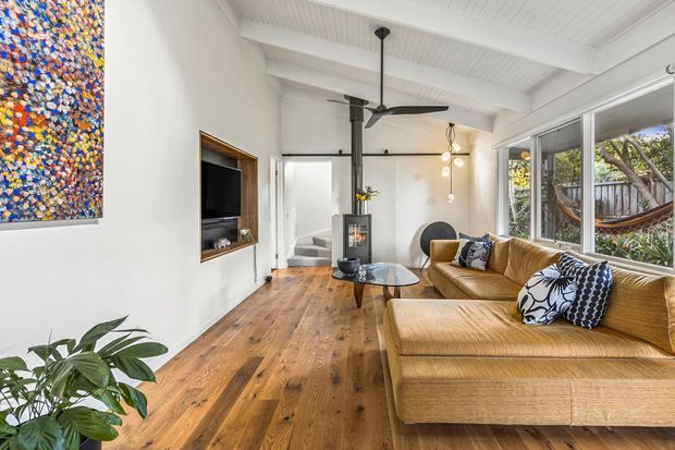 A bright, open-plan living room with warm timber flooring, white walls, exposed white ceiling beams and a large corner window. The room features a mustard-gold sectional sofa, black-and-white patterned cushions, a low glass coffee table, a suspended black wood-burning stove, ceiling fan, recessed television niche, sculptural pendant lighting and a colourful abstract artwork. Trees and a hammock are visible outside the glazing.