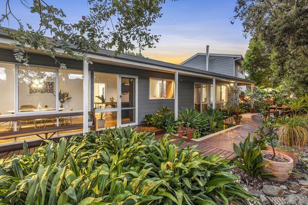 Exterior view of a single-storey contemporary home surrounded by dense landscaped planting. The dark grey weatherboard-clad facade has extensive glazing, a covered deck or veranda with outdoor dining, and a timber boardwalk leading through the garden. A pitched metal roof and warm interior lighting are visible at dusk.