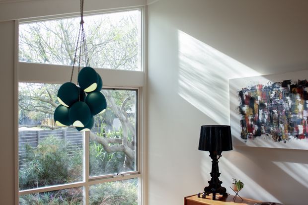 Bright contemporary interior corner with tall floor-to-ceiling windows overlooking landscaped greenery. A sculptural dark teal pendant light hangs in front of the glazing, while sunlight casts strong diagonal shadows across the off-white wall. A black table lamp and abstract multicolour artwork sit on a console, creating a refined, art-focused living space.