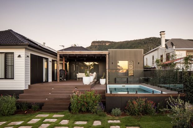 A contemporary residential backyard courtyard with a compact swimming pool, broad timber decking and steps, landscaped garden beds, and a covered outdoor entertaining pavilion. The home combines light horizontal cladding, dark tiled roofs, black-framed openings, and a corrugated metal-clad pavilion. A wooded rocky escarpment is visible beyond the property.