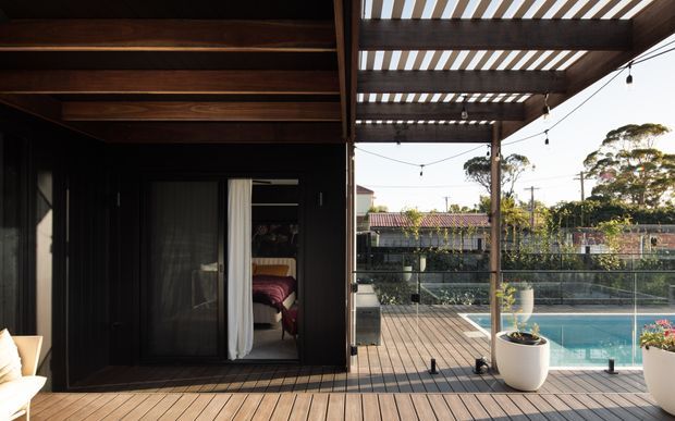 A timber-clad outdoor deck and covered pergola beside a swimming pool. The dark exterior wall includes a doorway revealing a bedroom beyond, while the deck has overhead timber battens, festoon lighting, large planters, and views toward neighbouring homes and trees.