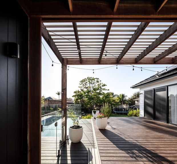 A spacious timber-decked outdoor terrace viewed from beneath a covered pergola. The pergola has exposed dark timber beams and slatted roofing, with festoon lights overhead. Glass balustrading borders a swimming pool, while large white planters, garden planting and a contemporary house facade with dark-framed glazing complete the scene.