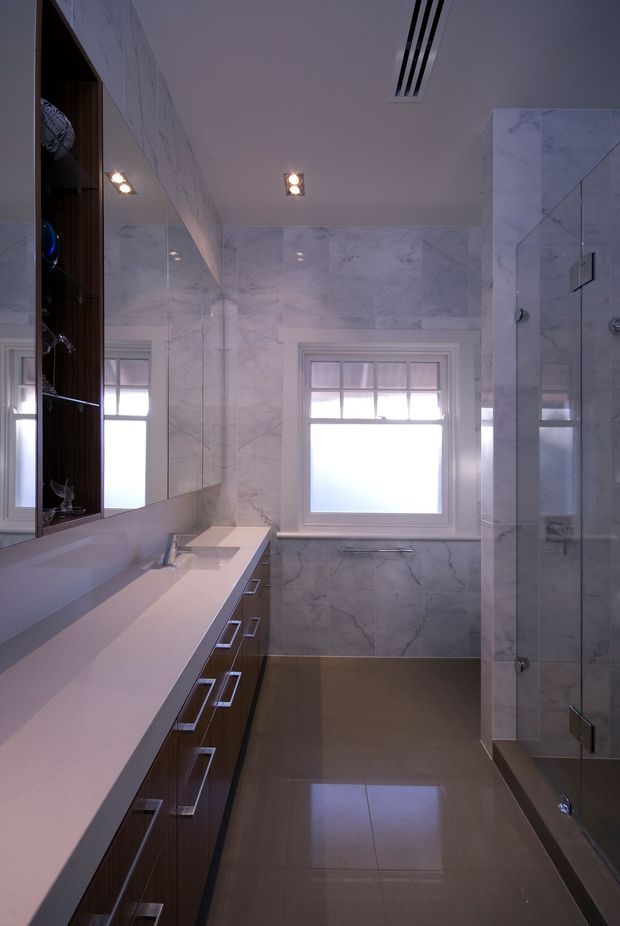 A refined contemporary bathroom with a long double vanity, dark timber cabinetry, pale engineered or natural stone benchtop, large-format marble-look wall tiles, and a fully glazed walk-in shower. Mirrored cabinetry and recessed ceiling lighting create a bright, polished space, while the glossy taupe floor adds warmth and reflectivity.