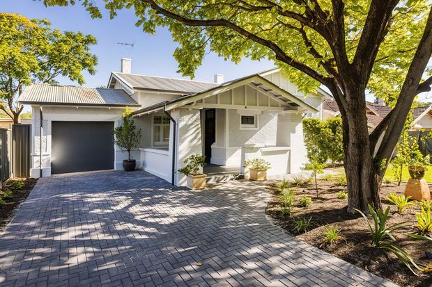 Street-facing view of a single-storey light-coloured bungalow with a prominent gabled porch, white rendered or painted masonry walls, corrugated metal roof, dark garage door, and paved brick driveway. Mature trees, clipped hedges, and landscaped garden beds frame the front facade.