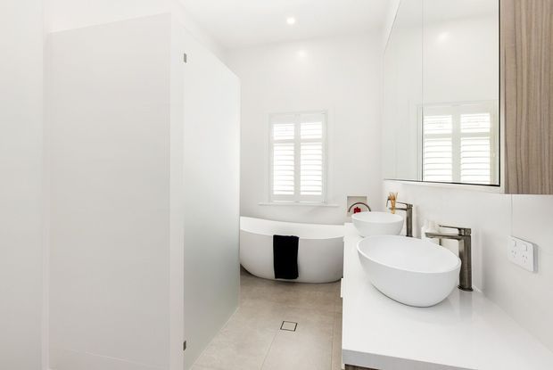 Bright contemporary bathroom with a freestanding white oval bathtub, double vessel basins on a white vanity, large wall mirror, pale tiled floor, white walls, and shuttered windows. A partial wood-grain storage cabinet is visible at the right.
