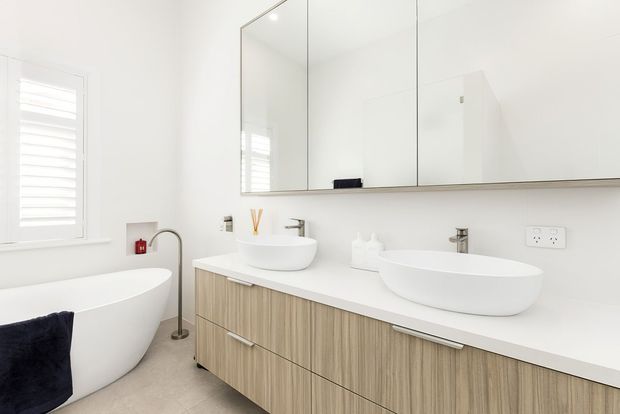 Bright contemporary bathroom with a freestanding white bathtub, double vessel basins, a long floating timber-look vanity, wall-mounted mirrored cabinetry, and a white plantation-shutter window. The space has a clean, minimalist fit-out with pale neutral finishes and abundant natural light.