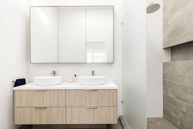 Bright contemporary bathroom featuring a double wall-mounted vanity with two white vessel basins, mirrored wall cabinetry, pale timber-grain joinery, and a walk-in shower lined with large-format grey-beige tiles.