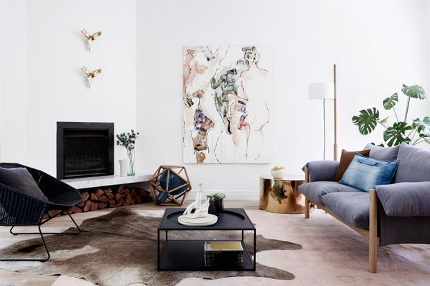 A bright, refined living room with a minimalist black fireplace set into a white wall, stacked firewood beneath, a low black metal coffee table, sculptural lounge chairs, and a large blue-grey sofa. Botanical plants, contemporary artwork, a slim floor lamp, and a cowhide rug add organic and artistic accents.
