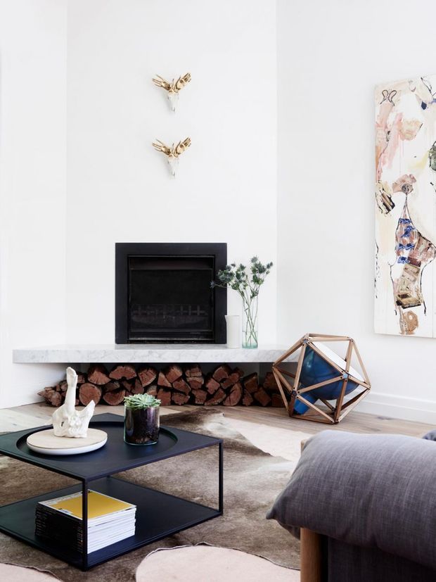 Bright, refined living room centred on a black built-in fireplace with a pale stone mantel and an open log storage recess beneath. White walls, sculptural brass wall accents, abstract artwork, a low black coffee table, neutral rug, and a large grey upholstered sofa create a clean contemporary interior with warm natural details.