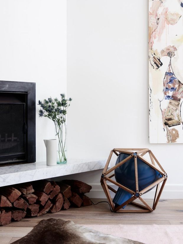 A close-up interior vignette of a contemporary fireplace with a dark stone surround and black firebox. A white marble hearth shelf holds a simple white vase and a glass vase with greenery, while stacked firewood fills the open space beneath. Beside the fireplace is a sculptural geometric timber-and-blue-leather chair or side piece. A pale abstract artwork hangs on the white wall, and light timber flooring with a pale rug completes the scene.