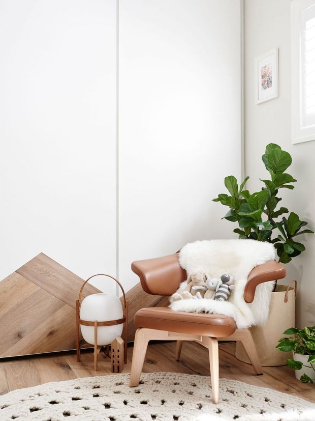 Bright, softly styled reading corner with a cognac leather lounge chair and matching ottoman, layered with a white sheepskin throw and plush toys. A large leafy indoor plant, pale neutral walls, white plantation shutters, framed artwork, a sculptural floor lamp, and warm timber flooring create a calm, Scandinavian-influenced contemporary interior.