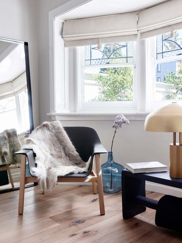 Bright, thoughtfully styled sitting area with a black mid-century lounge chair draped in a sheepskin throw, set beside a dark low coffee table. White-framed windows with quilted Roman blinds provide abundant daylight, while a large black-framed mirror reflects the room. The space features pale timber flooring, a blue glass vase with flowers, and a sculptural table lamp, creating a calm contemporary mid-century-inspired interior.